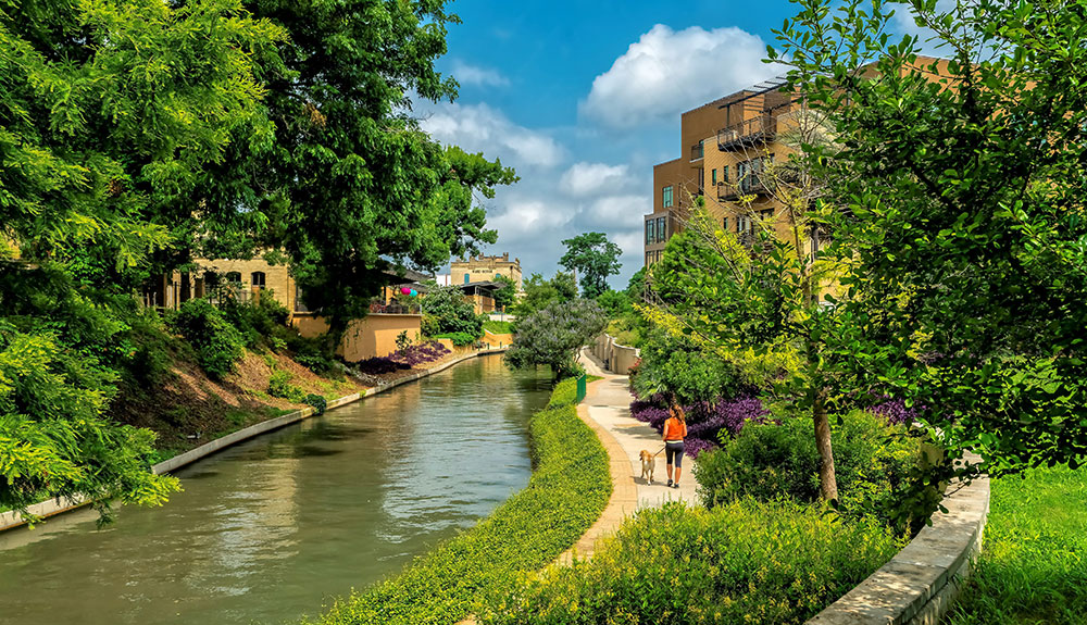 The river with cute buildings, trees, nature and a path with a person walking their dog on the famous River Walk in San Antonio, Texas