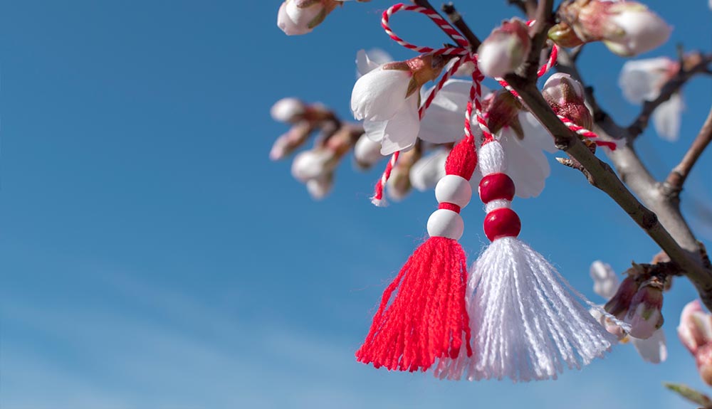 A red and white embroidery string bracelet is shown hanging from a tree with white blossoms on it