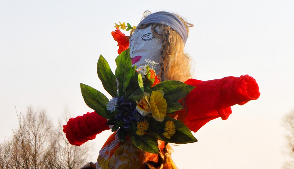 A puppet made of leaves and sticks with the painted face of a woman is shown outdoors