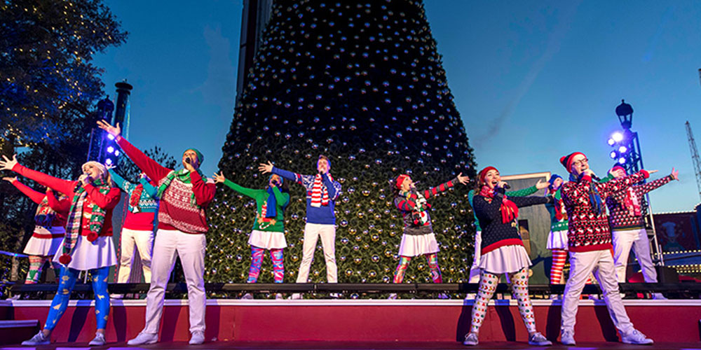 A christmas-themed theatre production; people stand on a red stage with a green christmas tree behind them, some are dressed in festive colours and others are wearing costumes like a nutcracker or a gingerbread man.