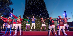 A christmas-themed theatre production; people stand on a red stage with a green christmas tree behind them, some are dressed in festive colours and others are wearing costumes like a nutcracker or a gingerbread man.