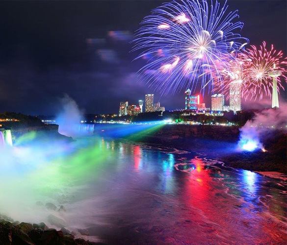 A waterfall at night is illuminated by rainbow-cololured spotlights is on the left of the image, while on the right of the image, above land, there are multiple high-rise buildings behind fireworks.