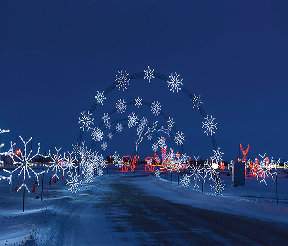 A paved, straight path going through a winter light show with arches above it covered in light up snowflakes. Real snow covers the ground on either side of the path.