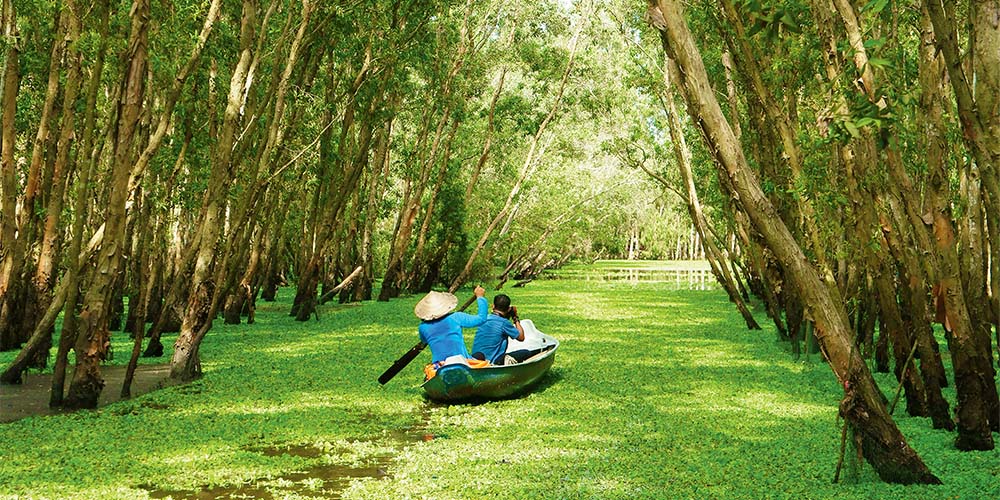 People wearing blue outfits on a small canoe going through a lake covered in leaves that have fallen on the river's surface from the trees that line the sides. The person holding an oar is wearing a beige straw hat.