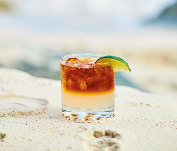 A cocktail in a short, clear glass with a lime on the rim on sand.