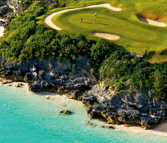 An overhead view of a cliffside golf course. The grass on the course is bright green, and goes to the edge of a rocky cliff. At the bottom of the cliff is a beach with clear, teal water.