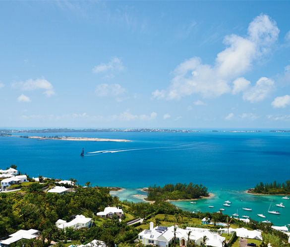 A view off the coast of Bermuda. The land leading up to the water is covered in green trees and white buildings, and the water closest to the short is a bright teal colour.