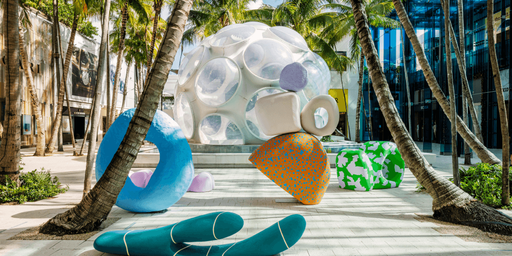 An outdoor foyer of a hotel; there are palm trees planted throughout the space and in the middle is a large water fountain and art piece. There are blue and green pool floaties on the ground in front of the water fountain.