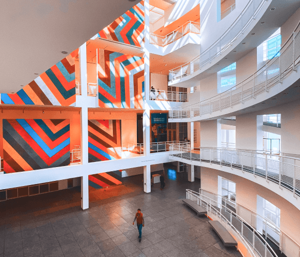 The inside of a building with an accent wall that is orange with stripes of blue and red, and white pillars. On the right side, the walls are white and there are doorways on each level.