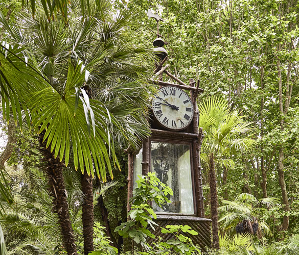 A tall, wooden clock in the middle of a garden, surrounded by trees and greenery.