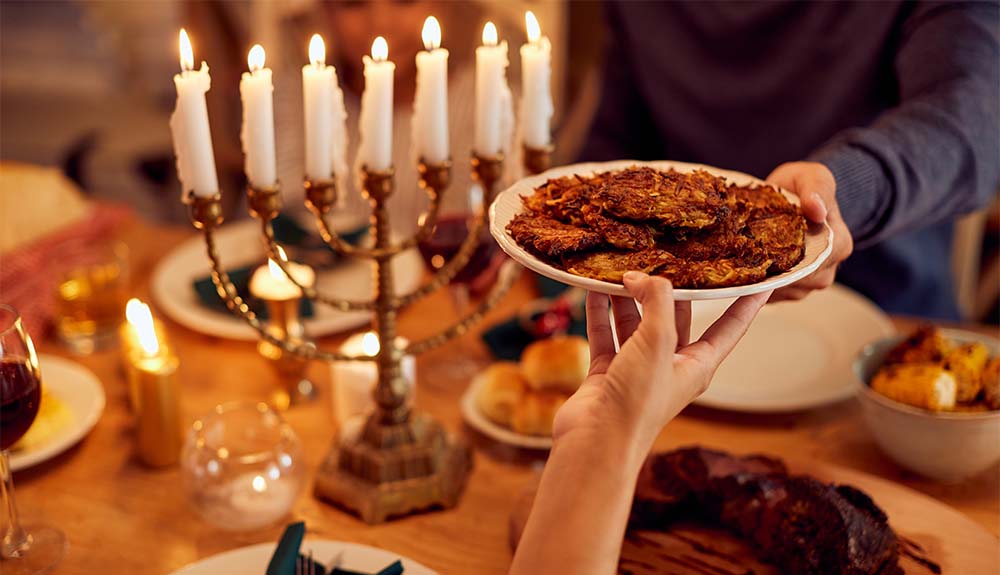 View of a table set up for a meal. There is a gold menorah in the background with candles lit. A person's hand is visible, holding up a plate of latkes and handing it to another person.