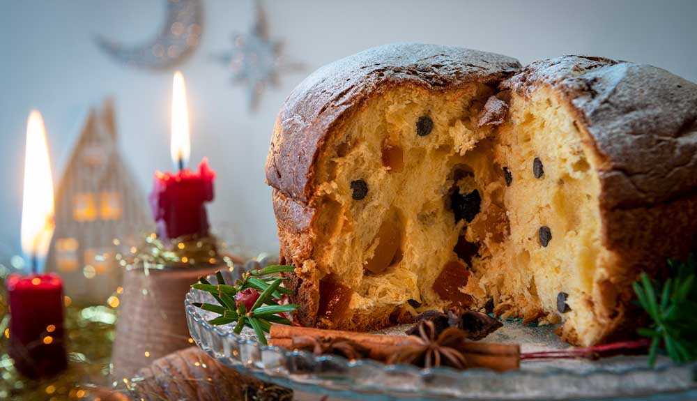 A golden yellow loaf of panettone is sitting on a clear glass cake stand with some cinnamon sticks and star anise on it. There are two lit red candles next to the cake stand.