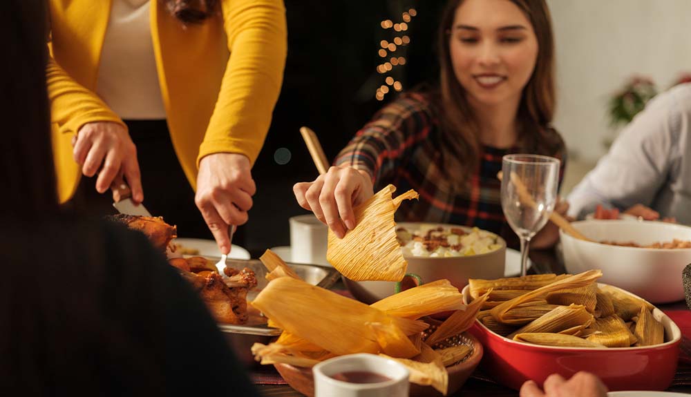Several people are sitting around a table. There is a person who is standing wearing an ivory blouse and a mustard yellow jacket. Next to that person is a woman seated, wearing a patterned shirt. She is holding a tamale wrapper in her hand.