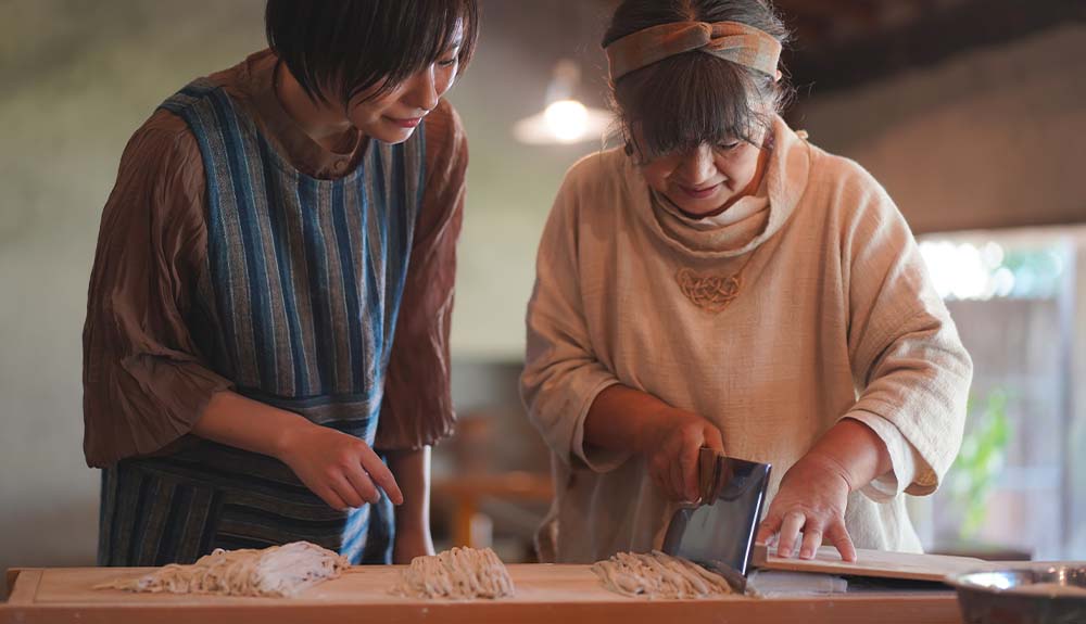 Two women, one older and one younger, are standing side by side at a cutting board. The older woman is holding a shiny metal in her hand, slicing some dough into long strips.