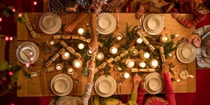 An overhead view of a table that's set up for a holiday meal. There are eight people around the table. There is some green pine branches set on the table with candles nestled in between the branches. There are also some holiday crackers on the table. Two people sitting across from each other are each holding onto one end of a holiday cracker.