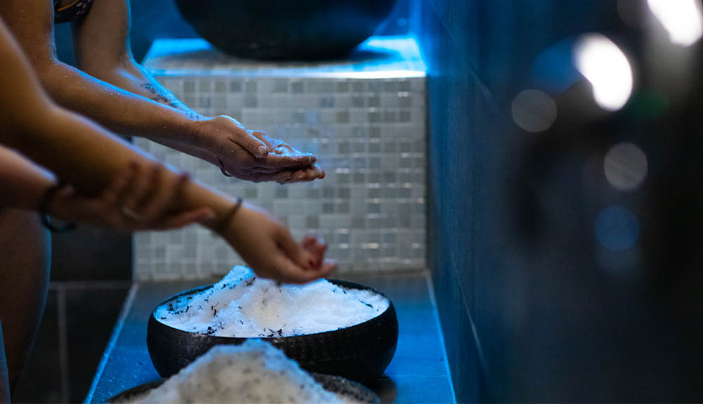 Two pairs of hands are hunched over large bowls overflowing with a white scrub. The people are rubbing the scrub on their hands and their arms.