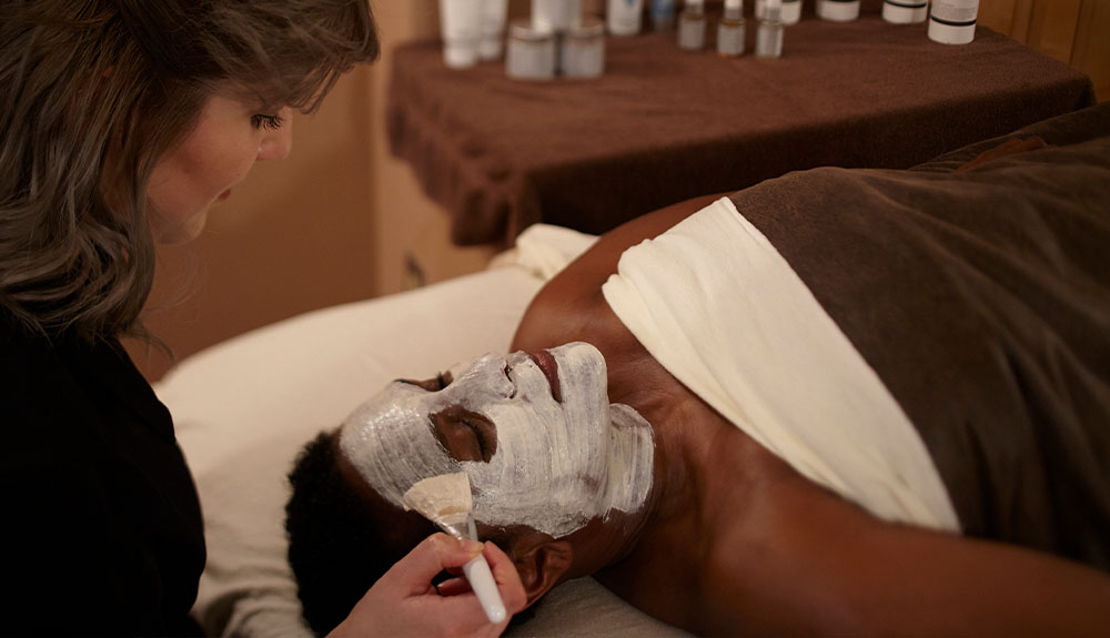 A sideview of a woman who is bent over a person lying on a bed, with blankets draped over their body. The woman is painting a white mask on their face with a paint brush.