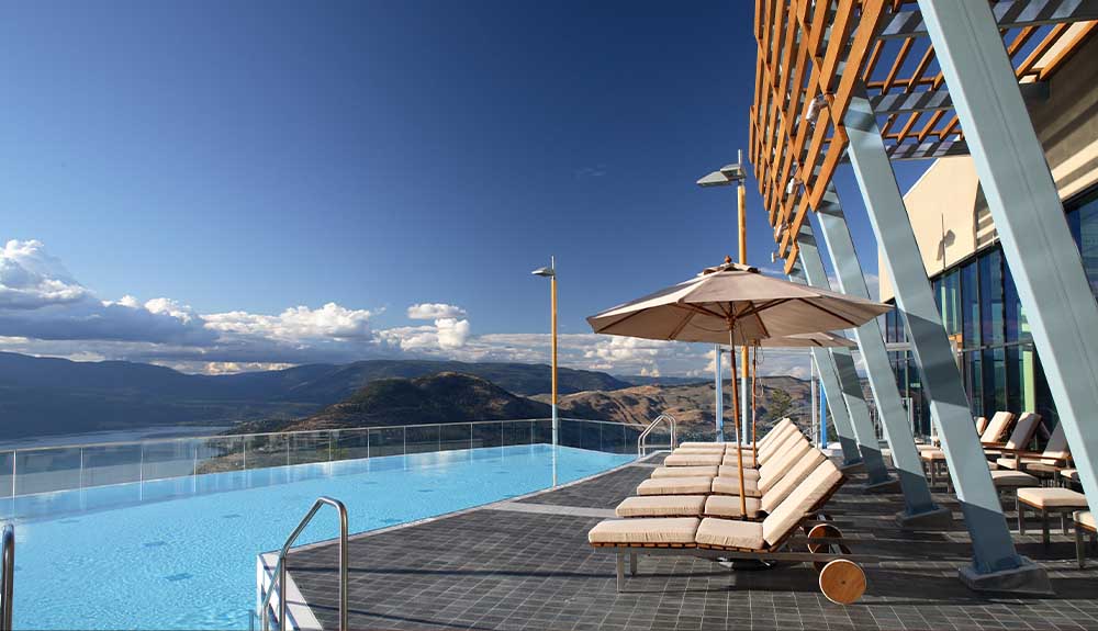 A row of ivory-coloured lounge chairs on a tiled deck facing an infinity pool that faces mountains and bright blue sky.
