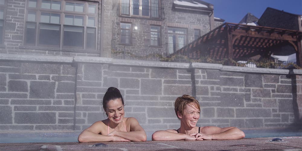 Two women are resting against the side of a pool. Behind them is a brick wall and a taller building looms behind them.