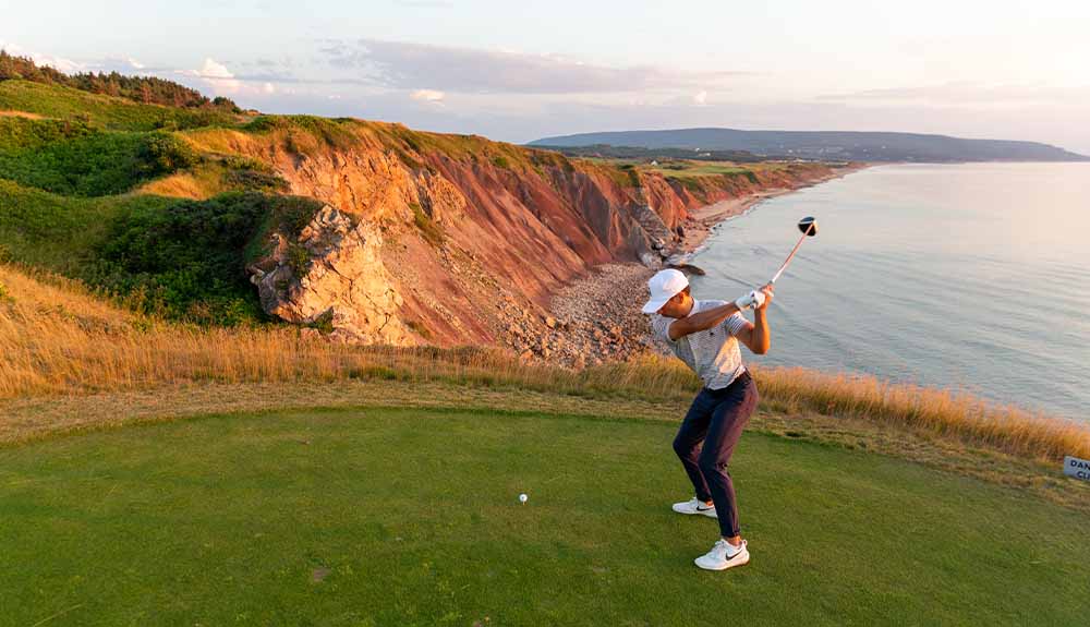 A lone golfer wearing dark pants and a white shirt, cap and shoes has his arms back, about to swing at a golf ball. Behind him is a body of water and cliffs.