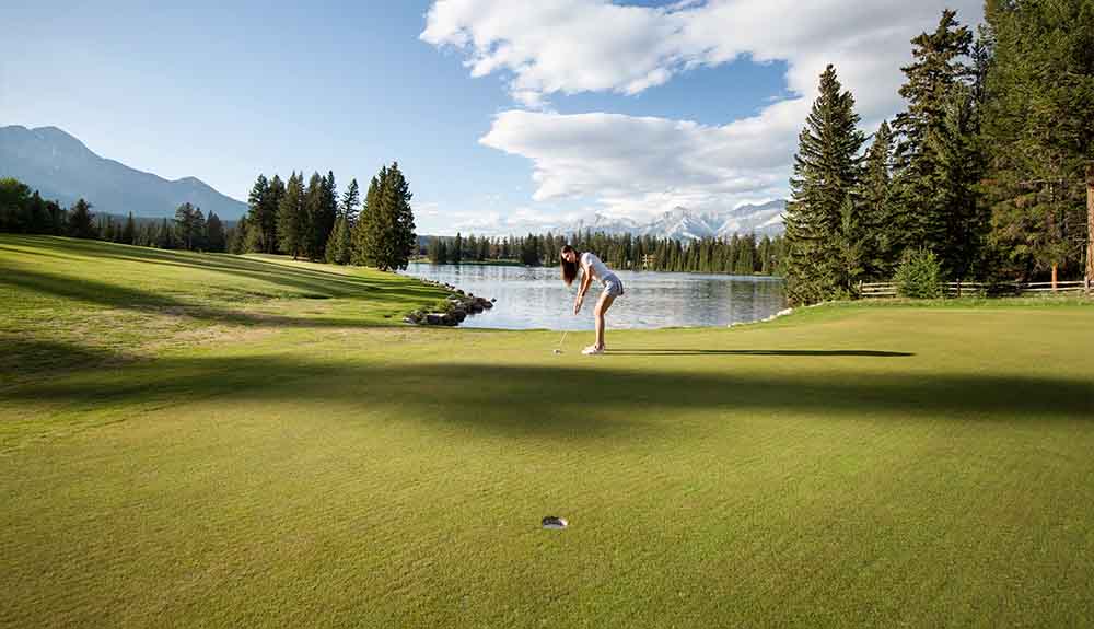 A female golfer with long brown hair wearing shorts and a T-shirt is bent over a club and about to hit a golf ball. Behind her are mountains and a sparkling lake.