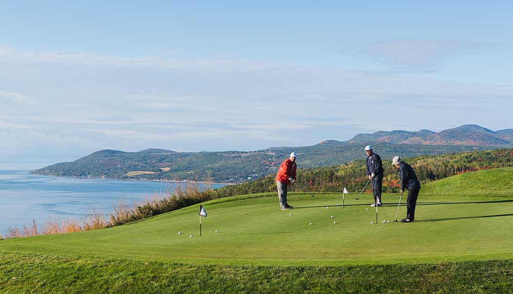 Three golfers are standing on a green course with mountains behind them and a lake on the other side. There are three hols and a series of golf balls scattered all over the green.