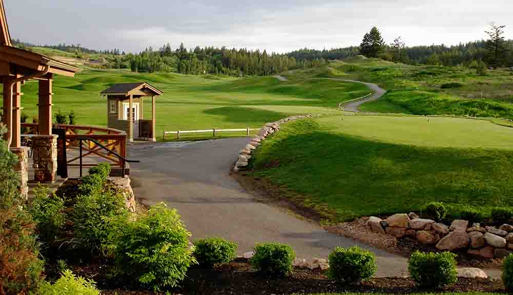 A rolling fairway with a dense row of trees in the background. There is a small wooden building with a white door and a small window off to the left hand side. There is a paved path down the centre of the rolling fairway.