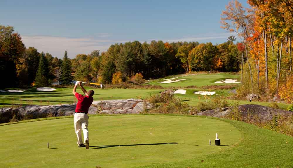 A golfer wearing a red polo shirt and khakis holding a club in the air after he's hit the ball over rolling greens with a dense forest of trees in the distance.