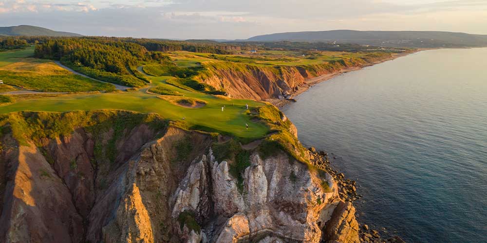 A rolling fairway on a cliff at sunset. The ocean is the right hand side and to the left are dense patches of trees.