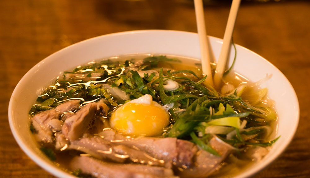 Bowl of soup with meat, egg and greens with a pair of chopsticks sticking out