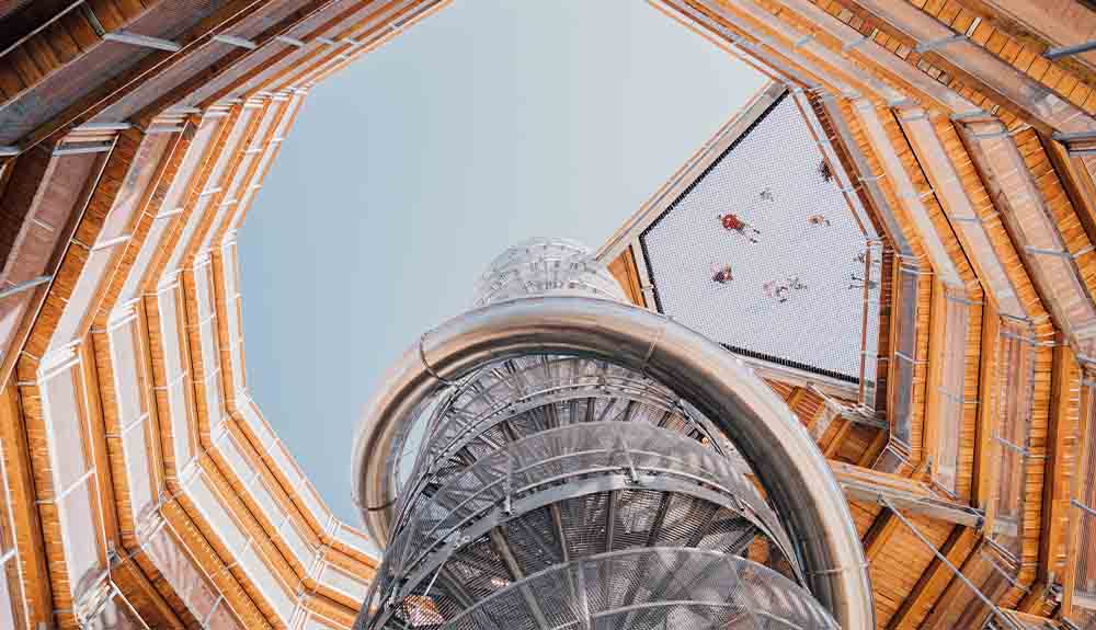 There is a silver spiral at the centre of the image. Surrounding is a wooden wall that opens up to the sky. There is a mirror or reflection at the top of the right hand side, where it’s possible to see a few people.