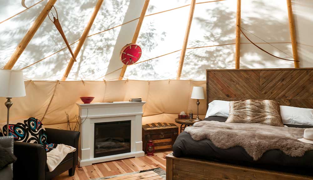 The inside of a domed space with a single double bed that is partially cut off. There is a hite fireplace on one side of the bed with a small red bowl on top of the mantle. There is a dark brown leather arm chair next to the fireplace with an Indigenous-print throw. On the other side of the fireplace is a small wooden chest. Next to it is a small round side table with a single lamp on it. 