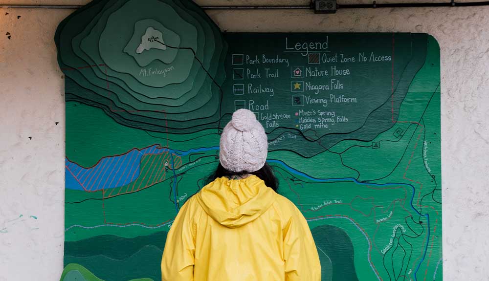 The back of a person wearing a bright yellow coat and a light grey knit toque with a pom pom is looking at a map. The map is predominantly green with Mt. Finlayson marked in the top left corner in a series of circles. There is a legend in the top right hand side with several places identified, including Nature House, Park Trail and Park Boundary. 