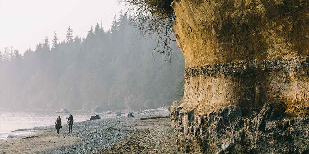 Two people are walking side by side on a rocky beach. It looks grey and overcast. Behind them are tall trees in shadow. To the right side, are rock formations with moss growing on them.