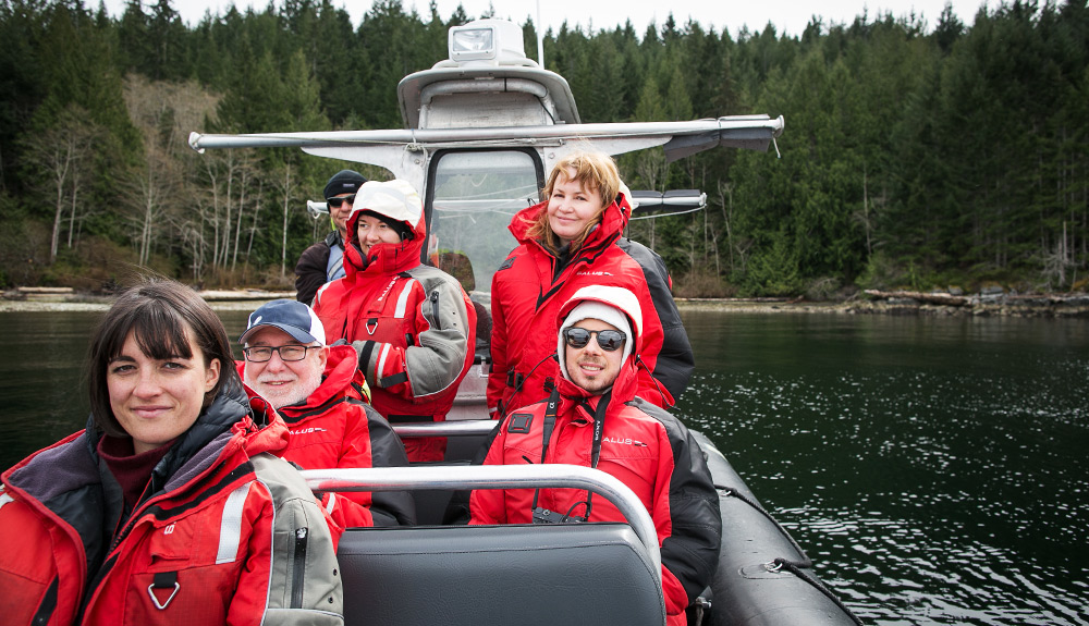 Five smiling people wearing thick red jackets sit in a pontoon boat, the captain wearing sunglasses and smiling behind them on a Zodiac ocean tour