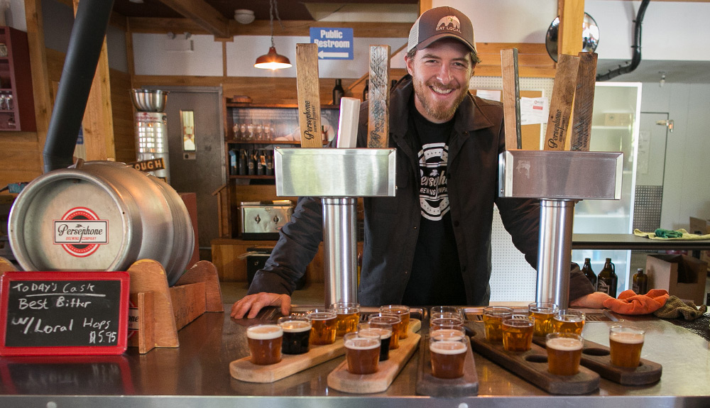 A young man in a baseball cap and blazer smiles from behind the bar at Gibsons Public Market, five flights of beers in front of him