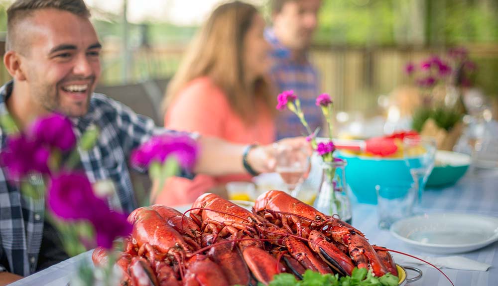 Three people sitting at a long dining table. Two of the people are out of focus. There are colourful serving bowls in front of them. In focus is a plate piled with freshly cooked lobsters.