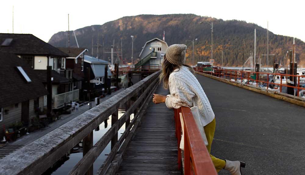 A woman wearing an off-white cableknit sweater, mustard coloured pants and a beige toque is standing at a bridge overlooking some houses. There are mountains behind her.
