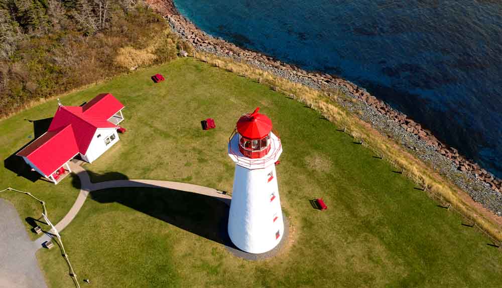 An aerial view of a a farm. There is a white building in one corner with a bright red roof and road ways coming out of it. There is a tall white light house with a bright red roof. On the other side is water.