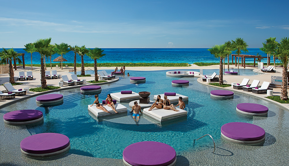 A large pool with purple circular pods and white square pods where several people lie socializing at the Breathless Riviera resort in Cancun, Mexico