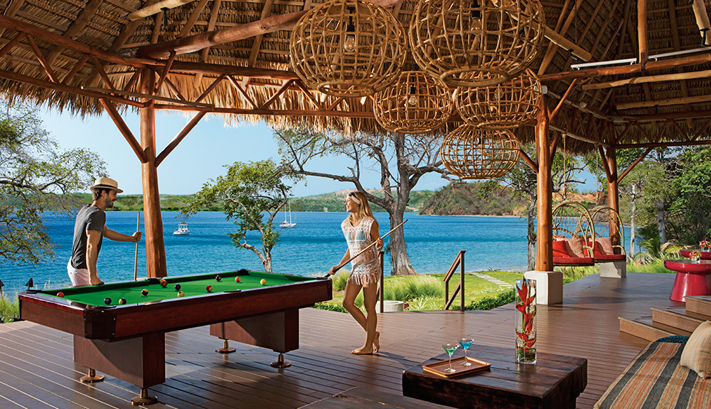 A man and woman in light summer clothes play a game of pool on the outdoor deck at Secrets Papagayo resort in Costa Rica