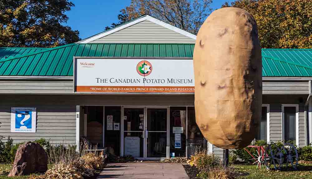 A wide building with a white sign that says The Canadian Potato Museum on it. At the forefront of the photo is a giant sculpture of a potato and behind it are some farming tools.