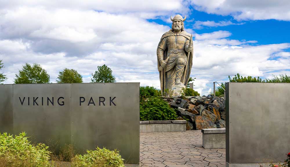 A metal sign to the left of the image says Viking Park in capital letters. Behind it is a life-size statue of a viking wearing a hat and a cape, holding a sword in one hand, with one knee raised on a rock.