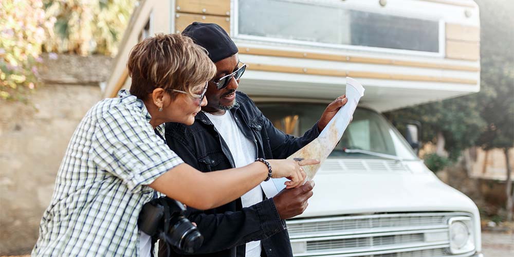 Two people, a man and a woman, are standing in front of an RV. The man is holding a map and the woman, who has a SLR camera around her neck, is looking over his shoulder and pointing at a spot on the map.