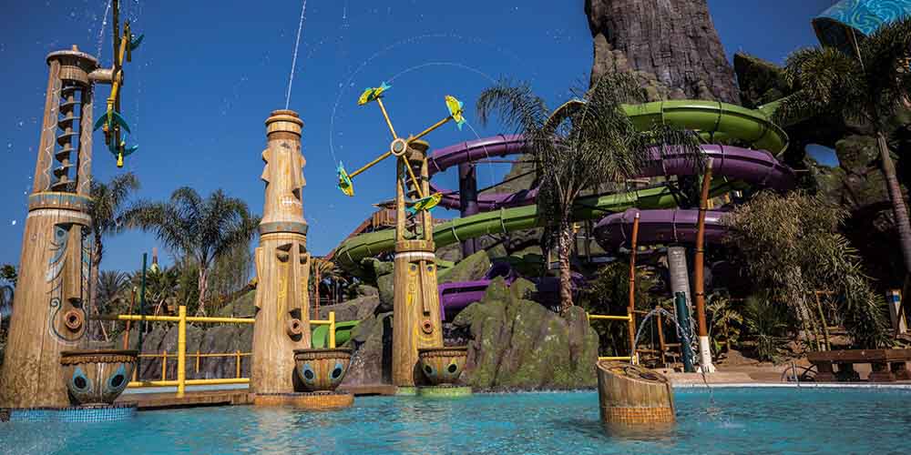 Outdoor water park with green and purple water slides winding around a wooden structure with wooden figures with faces spitting water from the their tops