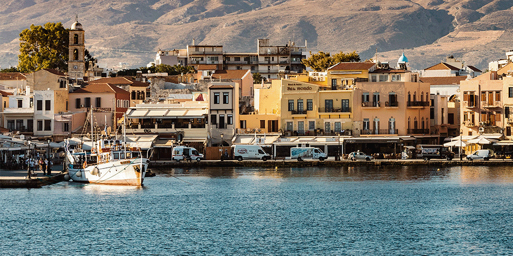 A landscape image of a town beside a dock. At the forefront of the image is calm, blue water. To the left there is a single white boat in the water and a few people standing on the dock near it. Lining the rest of the dock are crowded buildings in different shades of beige, peach and yellow, and on the street in front of them are white vans. Behind the buildings are steep, brown hills.