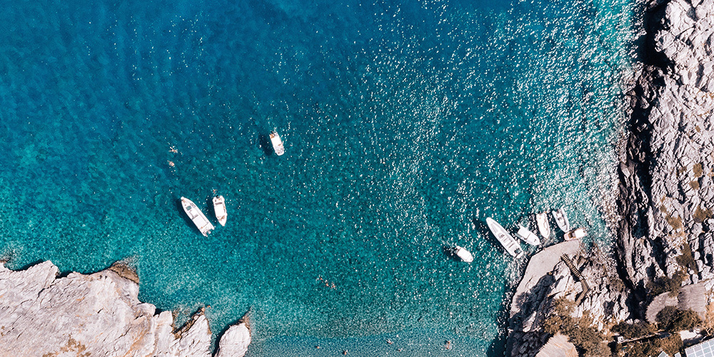 An overhead shot of a rocky shore and shallow turquoise water. There are 9 small boats scattered in the water near the grey shore. The sun is shining brightly and reflecting off of the water.