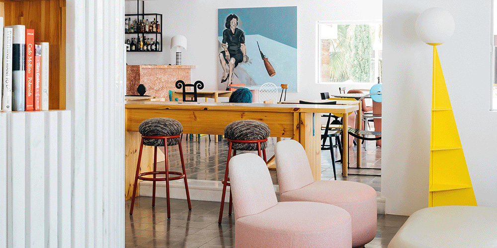 An interior shot of a hotel lobby. There’s a large window at the back of the room, letting a lot of light in. Two pink, cushiony chairs are the forefront of the image, and behind those is a long wooden table and two stools with red frames and grey cushions. There is a painting on the back wall of a woman in front of a blue background, and there is a small bookshelf with a few books on the left side.