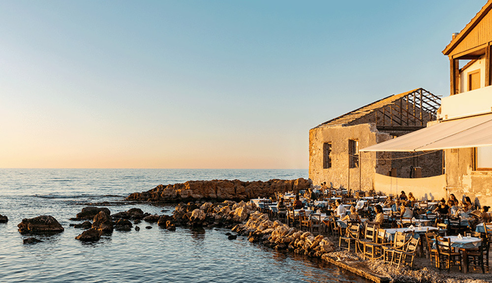 A landscape photo of a seaside restaurant. The sun is setting and the water is calm, with boulders leading up to the restaurant on the right side, half-filled with diners. Part of the restaurant’s patio is under the shade of a white tent. The chairs and tables are all wooden, and the tables are covered in white tablecloths. 