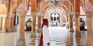 A man in a white robe and red turan walks through the beautiful pink stone colums in a building in Japur, India